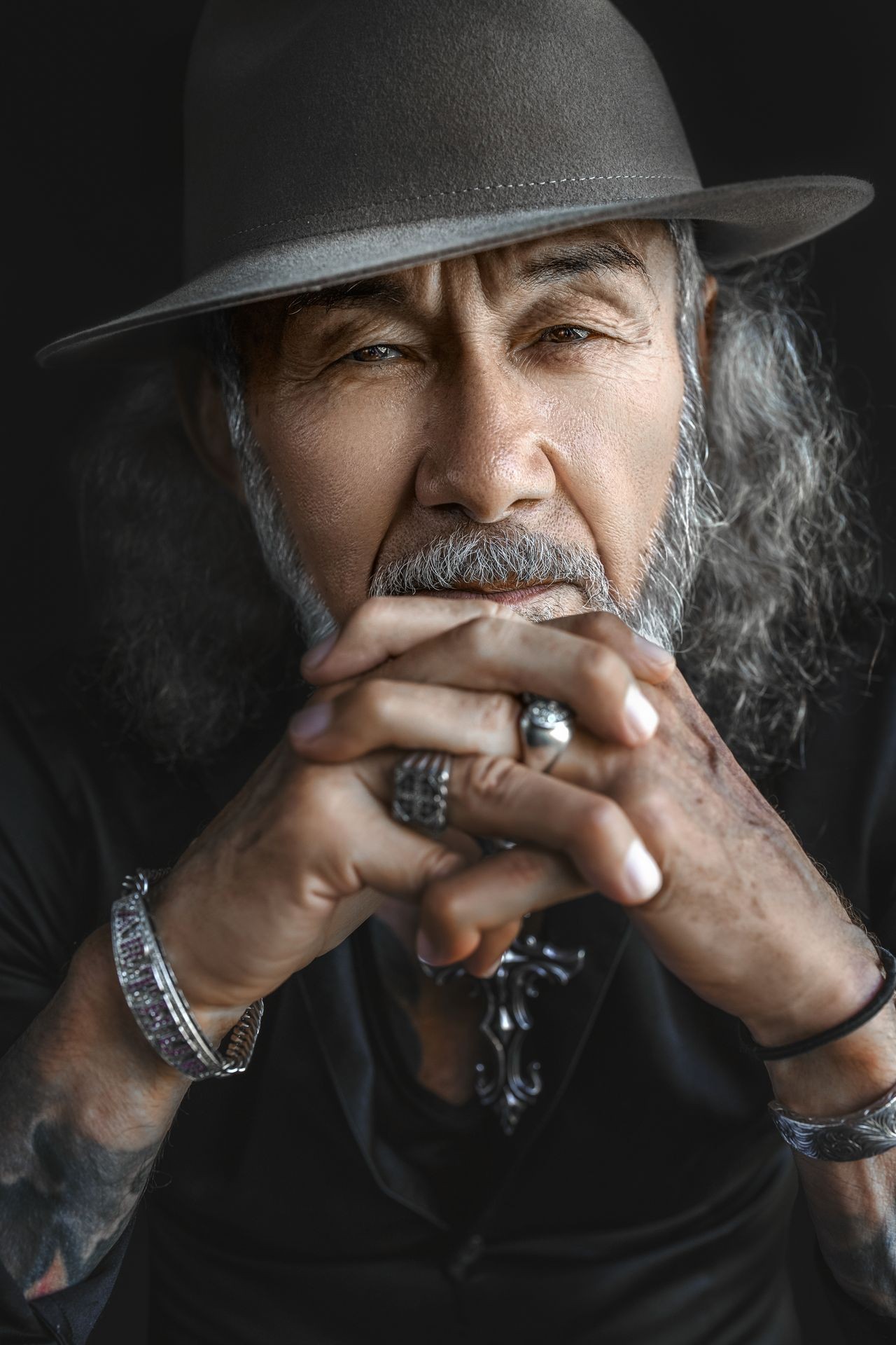 Elderly man wearing a hat, with fingers interlocked, showing rings and a bracelet.