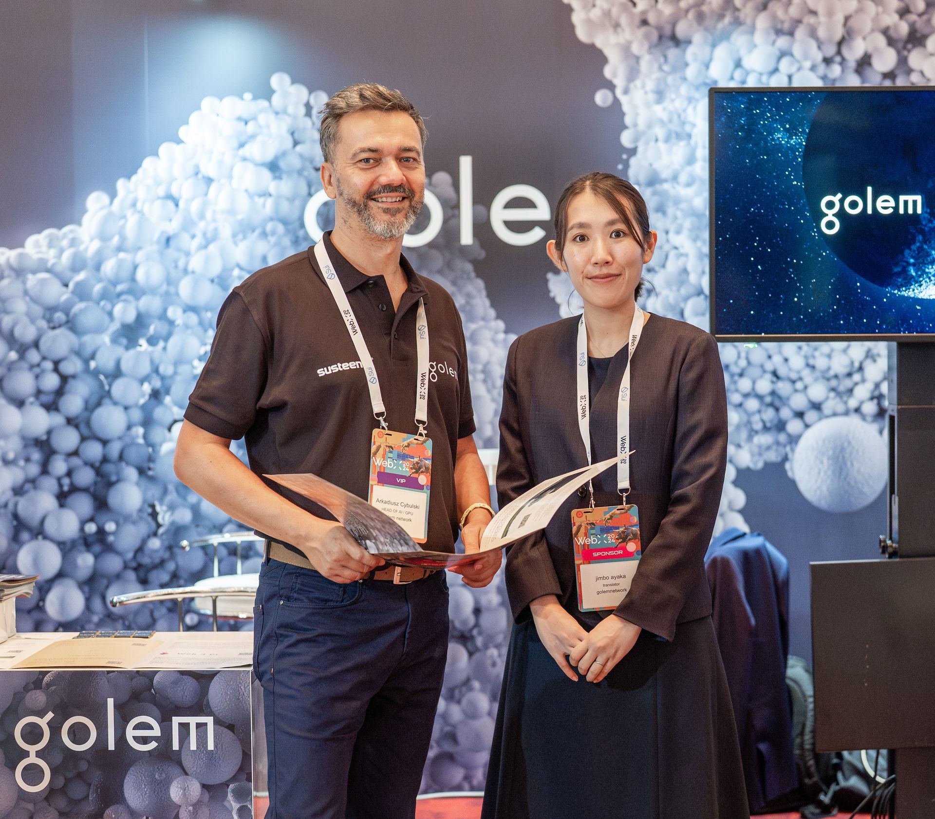 Two people standing at a booth with 'golem' logo in the background and holding brochures.